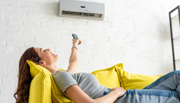 Eine junge Frau liegt entspannt auf einem leuchtend gelben Sofa und richtet eine Fernbedienung auf eine Klimaanlage an der Wand &uuml;ber ihr.