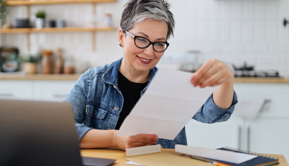 Eine Frau mit grauen Haaren und Brille sitzt in ihrer Küche und liest auf einem Blatt.