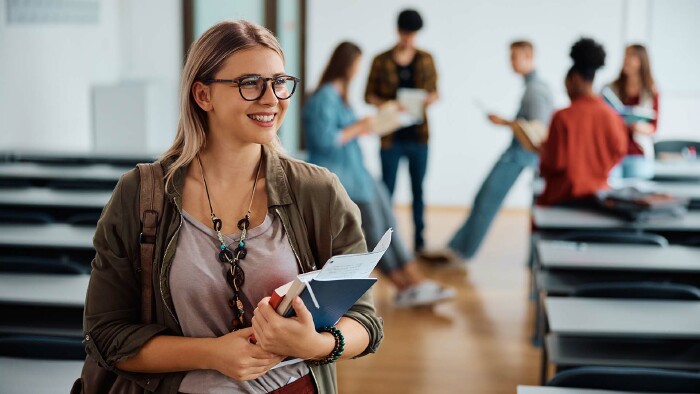 Junge Frau mit Brille steht l&auml;chelnd in einem Klassenraum und h&auml;lt Unterlagen in der Hand. Im Hintergrund unterhalten sich mehrere Studierende in kleinen Gruppen.