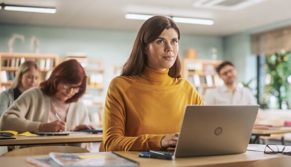 Eine Frau in einem gelben Rollkragenpullover sitzt in einem Klassenraum an einem Laptop. 
