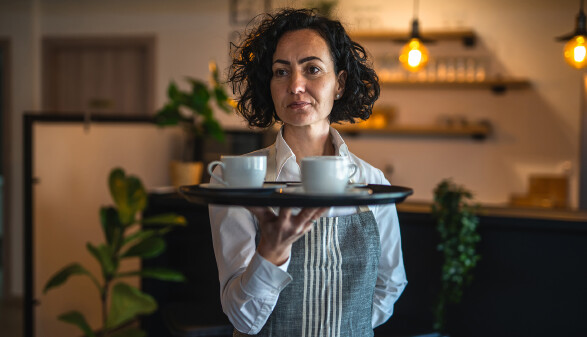 Kellnerin trägt Tablett mit zwei Kaffeetassen mit ernstem Blick durch ein Café