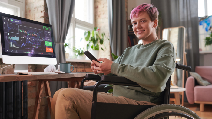 Junge Frau mit kurzen pinken Haaren sitzt mit Handy in der Hand im Rollstuhl vorm Computer, auf dem eine Seite mit Aktienkursen ge&ouml;ffnet ist. Sie l&auml;chelt in die Kamera.