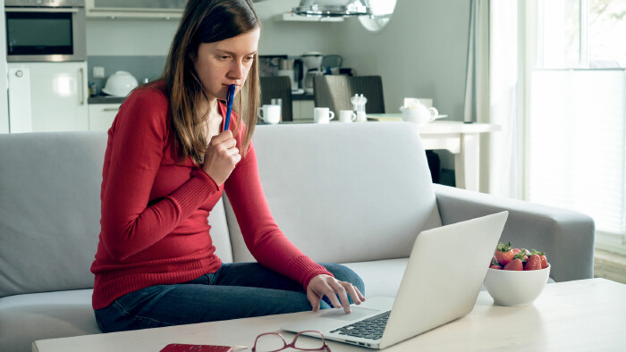 Frau sitzt auf Sofa, arbeitet konzentriert am Laptop, h&auml;lt Stift in der Hand, helle moderne Wohnung.