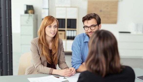 Junges Paar sitzt in einem B&uuml;ro an einem Tisch und spricht freundlich mit einer Beraterin, auf dem Tisch liegen Dokumente.